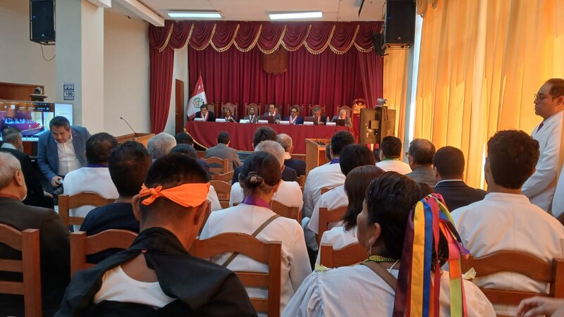 a packed courtroom seen from the back with dozens of people wearing colourful headwear; they are looking towards a stage area with a red curtain, in front of which a long table with 7 men and one woman sit in front of nameplates. There are screens, microphones and cameras pointing at the table and towards the seated audience.